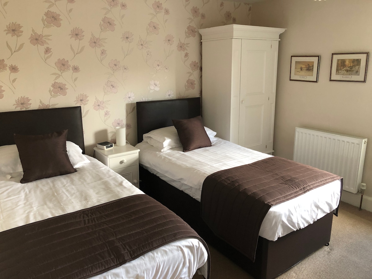 A serene guest room features two beds with dark brown headboards, complemented by elegant brown throws and coordinating cushions. Floral wallpaper in soft colors enhances the calming environment. A white nightstand and wardrobe provide practical storage, while framed art adds a touch of charm.