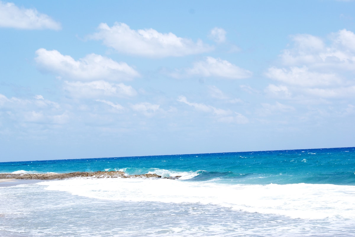 The image captures a serene ocean view featuring vibrant blue waters gently meeting a sandy shore. Waves create a frothy white crest as they roll onto the beach. A clear sky, dotted with soft clouds, extends above the horizon.