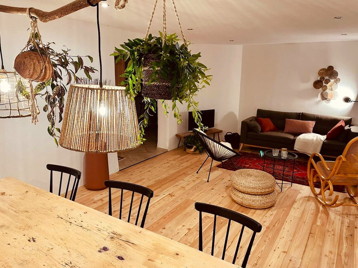 A welcoming open living space features wooden floors and is adorned with greenery from hanging plants. A large wooden dining table is complemented by black chairs. A cozy seating area is arranged with a dark sofa and a decorative rug, creating an inviting atmosphere.