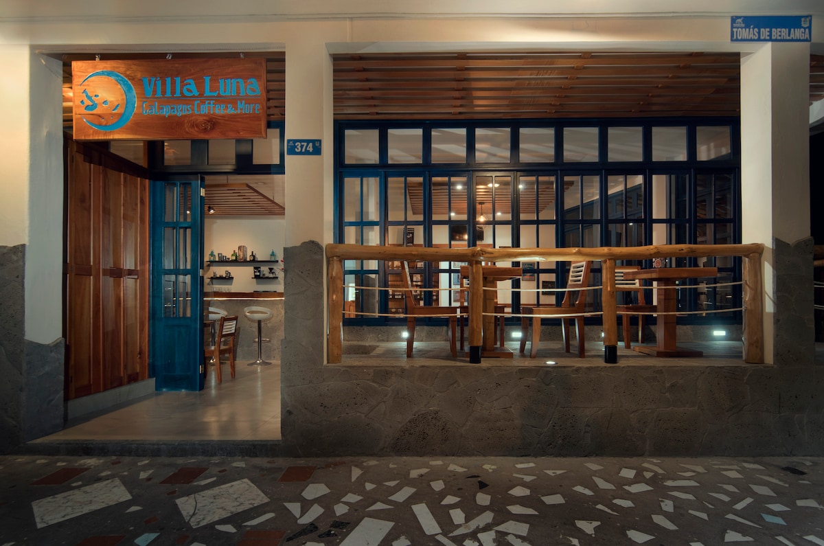 The entrance area of Villa Luna is captured, featuring a wooden railing and large glass doors framed in blue. The open space displays simple wooden tables and chairs, inviting guests to relax. Natural light filters in, creating a warm ambiance.