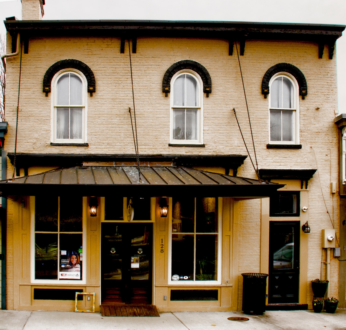 The historic building features arched windows on the second floor, accented by dark trim. A welcoming entrance is framed by large display windows, showcasing the business within. The awning provides shade above the entrance, and the exterior exhibits a neutral color palette.
