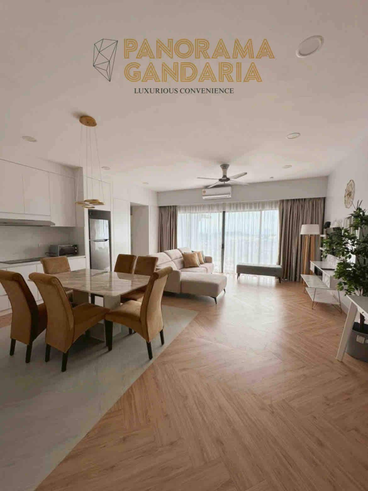 An open-concept living and dining area reveals a dining table set for six, accompanied by plush, tan chairs. A modern kitchen with sleek cabinetry is visible in the background. Natural light floods the space through large glass doors, leading to a balcony area with a view.