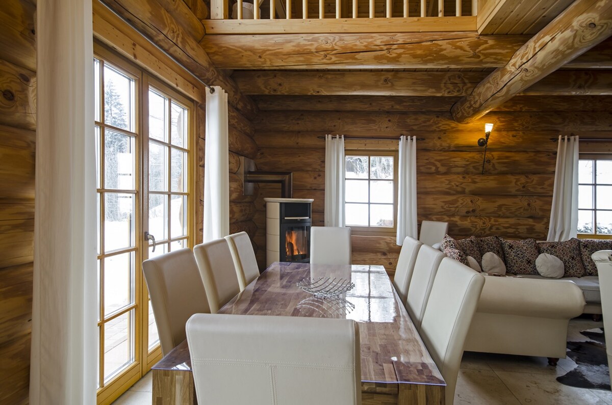 A spacious dining area features a long wooden table surrounded by light-colored chairs. Soft natural light enters through large windows, and a cozy seating area is visible in the background, offering a warm setting. Wooden beams and logs create a welcoming cabin aesthetic.