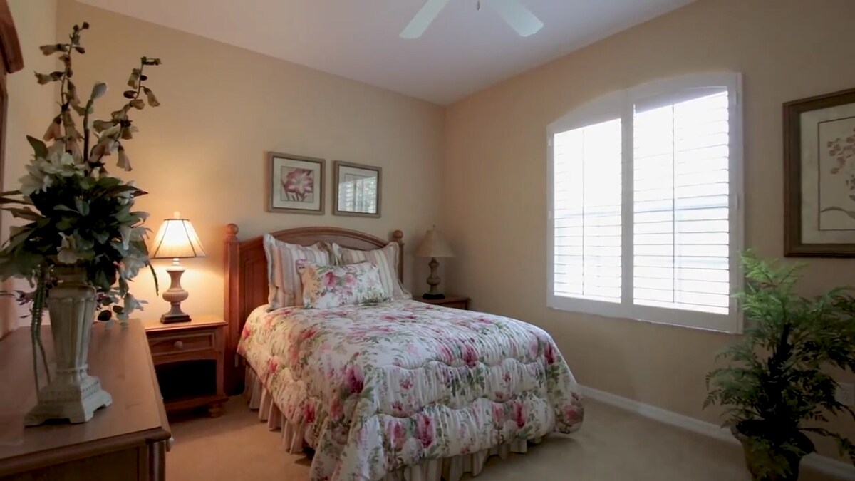 A serene bedroom features a floral-patterned bedding set on a spacious bed. Light-colored walls complement the wooden furniture, including a nightstand with a lamp. Natural light filters through the white, shuttered windows, and a decorative plant adds a touch of greenery.