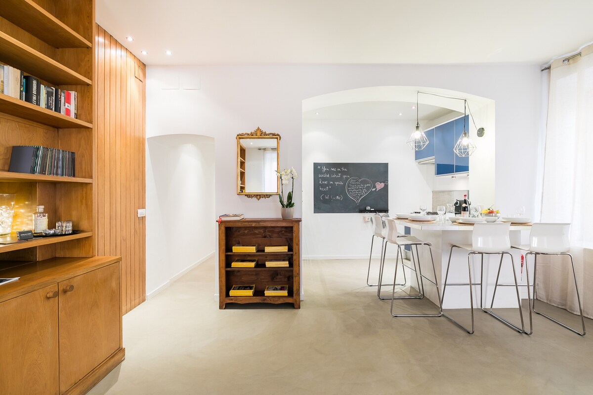A bright living space is displayed, featuring a stylish wooden bookshelf and a charming wooden cabinet. A modern kitchen island with high stools is positioned near a wall-mounted chalkboard. Natural light pours in through large windows, enhancing the open and airy atmosphere.