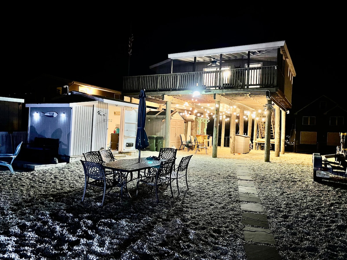 The exterior of the property is illuminated at night, featuring a raised deck with seating and a hot tub below. A dining set is positioned on the ground level, surrounded by neatly arranged outdoor furniture. Soft outdoor lighting enhances the inviting ambiance.