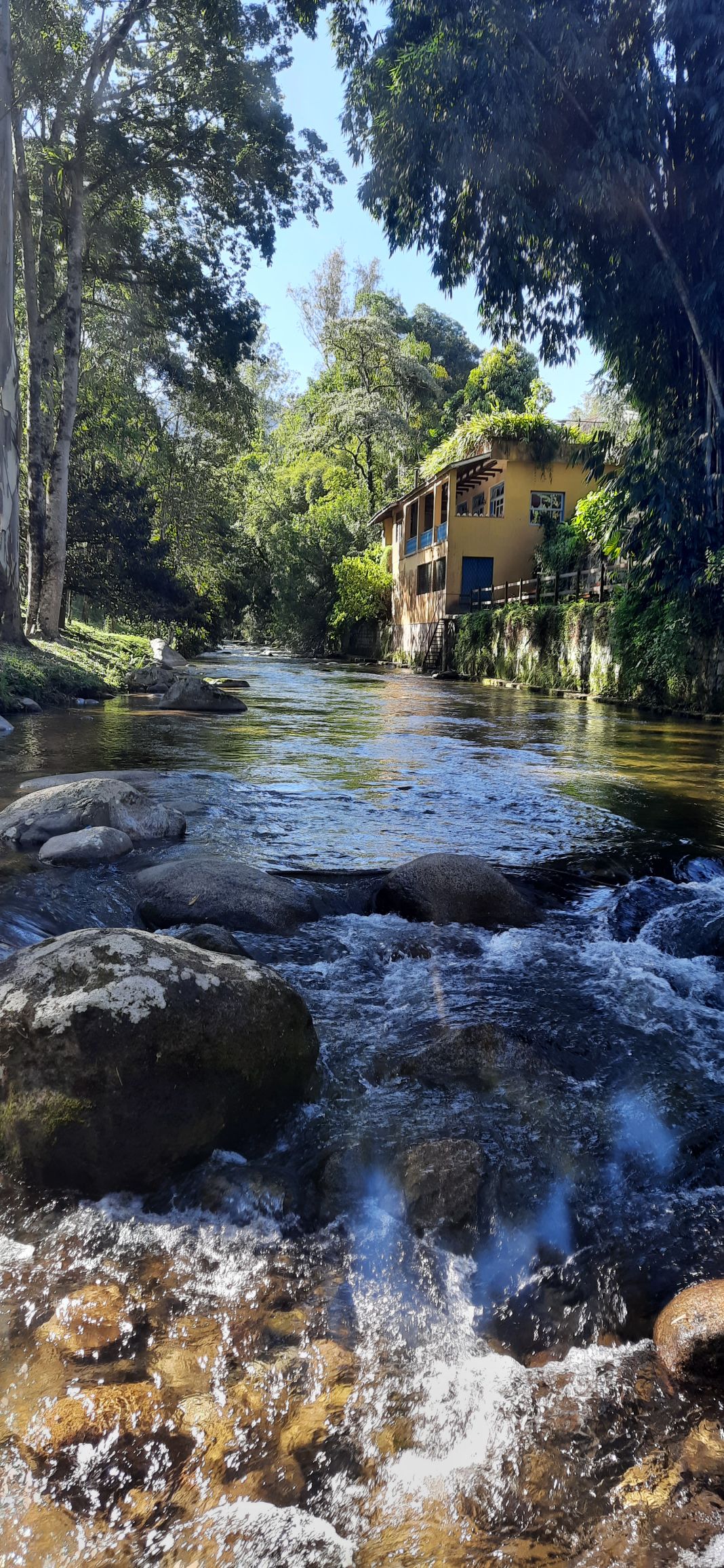 A serene view of a clear river flows gently over rocks, reflecting sunlight. On the right, a two-story chalet is nestled among green trees, showcasing a welcoming yellow exterior. The surrounding nature adds to the tranquil setting, enhancing the peaceful atmosphere.