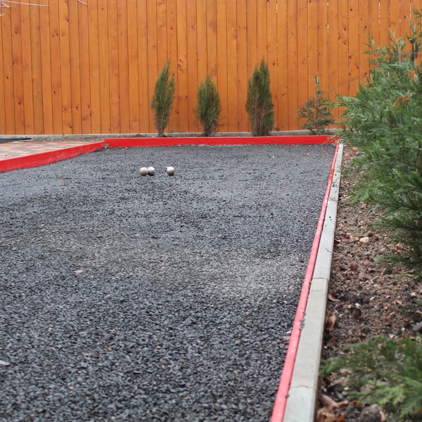 A petanque court is visible, featuring a gravel surface bordered by red edging. Two metallic balls are positioned on the court, with low greenery along both sides, creating a defined playing area.