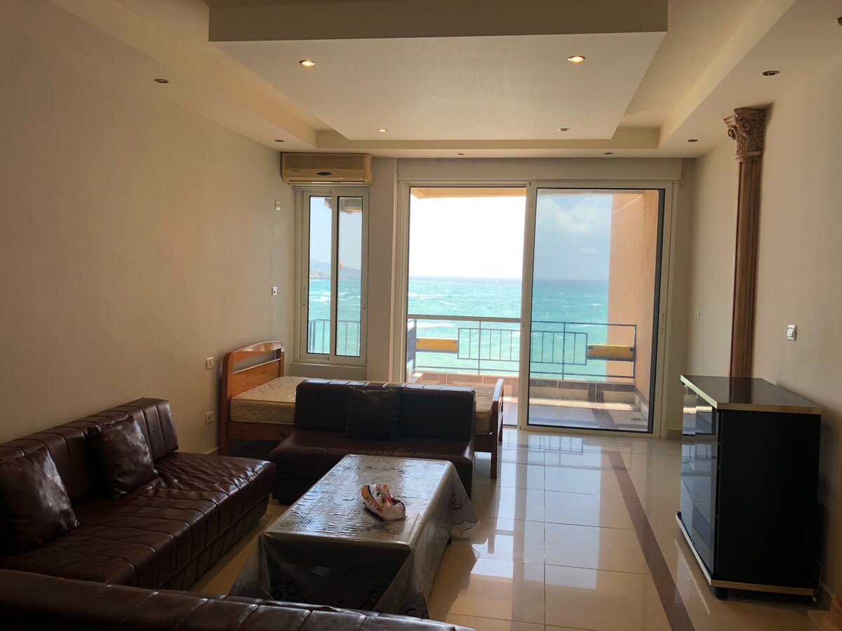 A spacious living area is showcased, featuring a large window that offers a direct view of the sea. Comfortable seating arrangements, including a dark leather sofa and coffee table, are present. A partially assembled bed can be seen in the background, along with a modern TV unit.