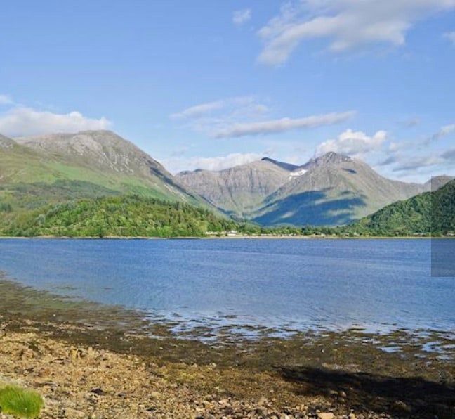 A serene landscape is depicted featuring a calm body of water bordered by rocky shores. Rolling hills and substantial mountains rise in the background, surrounded by lush greenery. The scene is brightened by clear blue skies with a few clouds drifting above.