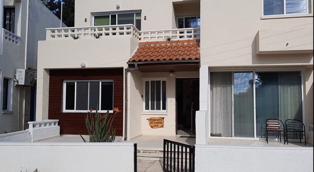 The villa's exterior features a charming entryway with a terracotta roof and a welcoming sign. Large windows provide ample natural light to the inviting entrance area. A small garden area is visible, with a mix of greenery and decorative elements.