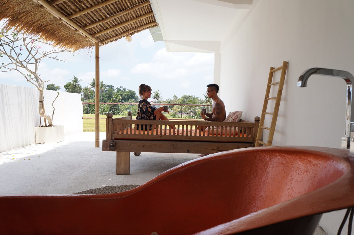 An outdoor private balcony features a natural wood seating area, where two guests are seated comfortably. An orange tub is positioned in the foreground, with a view of rice fields and gardens visible in the background, framed by a thatched roof above.
