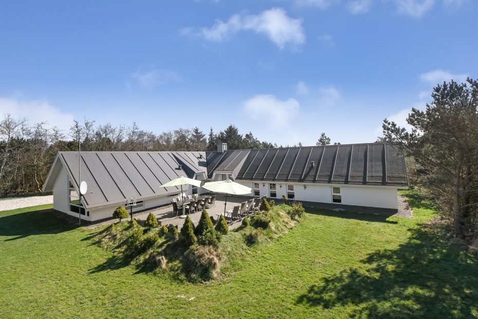 An expansive holiday home is set in a large grassy area, featuring a spacious patio with multiple seating arrangements. Umbrellas provide shade, while the building's modern design is highlighted by its sloped roof and multiple sections merging harmoniously with the natural surroundings.
