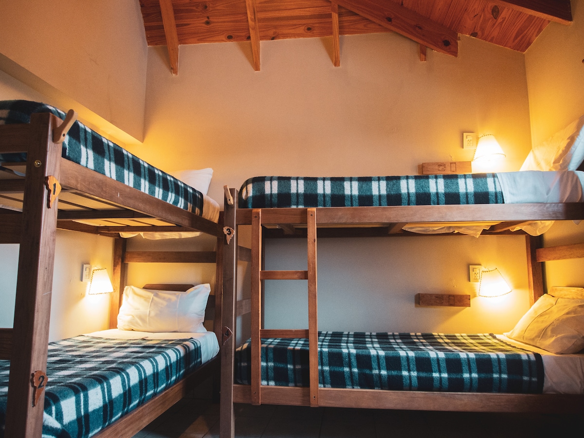 A cozy sleeping area features two wooden bunk beds, equipped with warm, checkered bedding. Soft wall-mounted lamps provide gentle lighting, complementing the wood-beamed ceiling. Spacious, light-colored walls enhance the room’s openness.