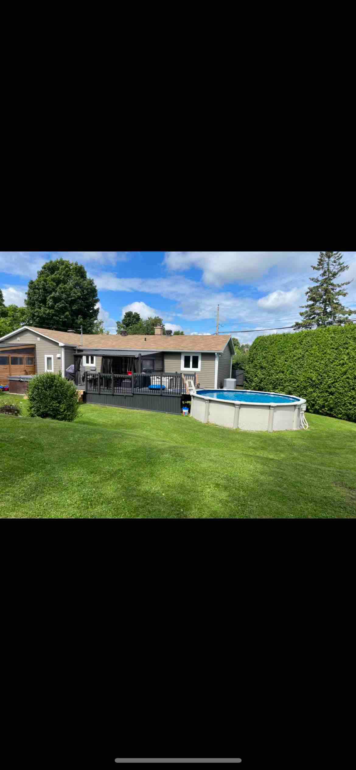 A well-maintained yard features a swimming pool and neatly trimmed hedges. The house, with a deck, provides easy access to outdoor spaces, where greenery surrounds the area. A clear sky with scattered clouds enhances the inviting outdoor setting.