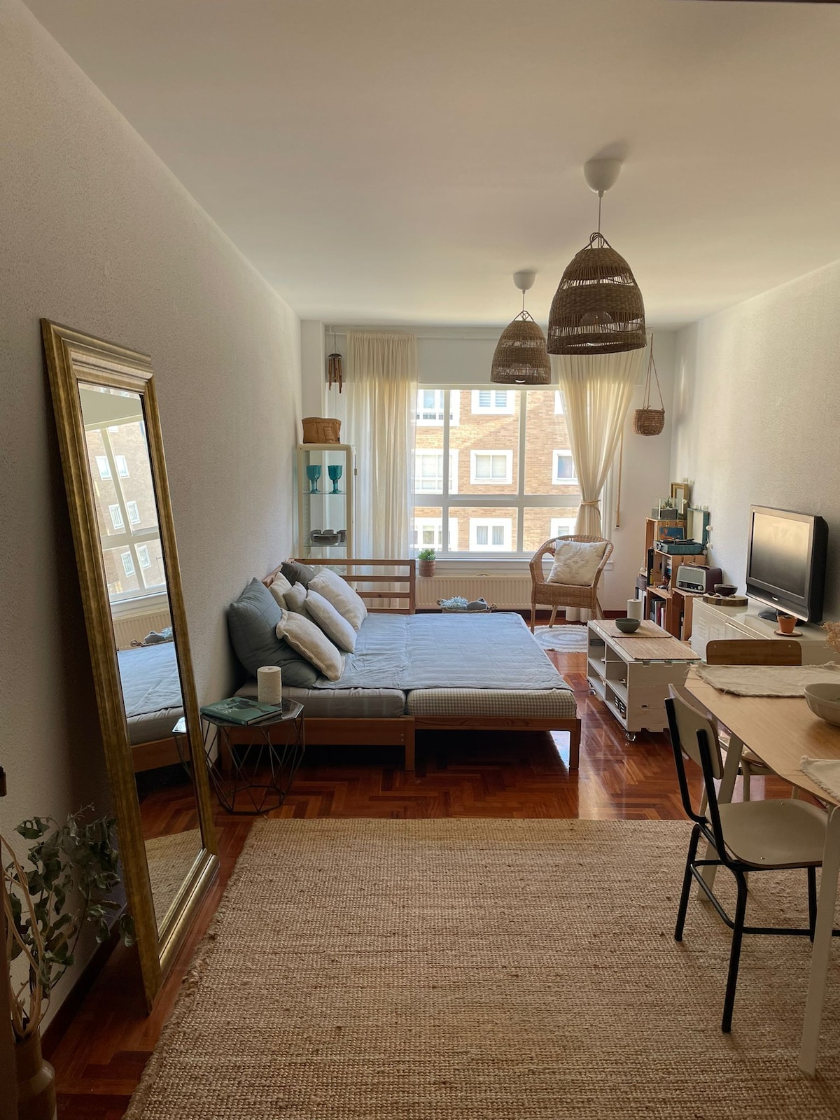 A spacious living area is furnished with a light grey sofa bed and a wooden coffee table. Natural light enters through large windows adorned with sheer curtains. A mirror reflects the room's features, while a dining area is set with a simple table and chairs.