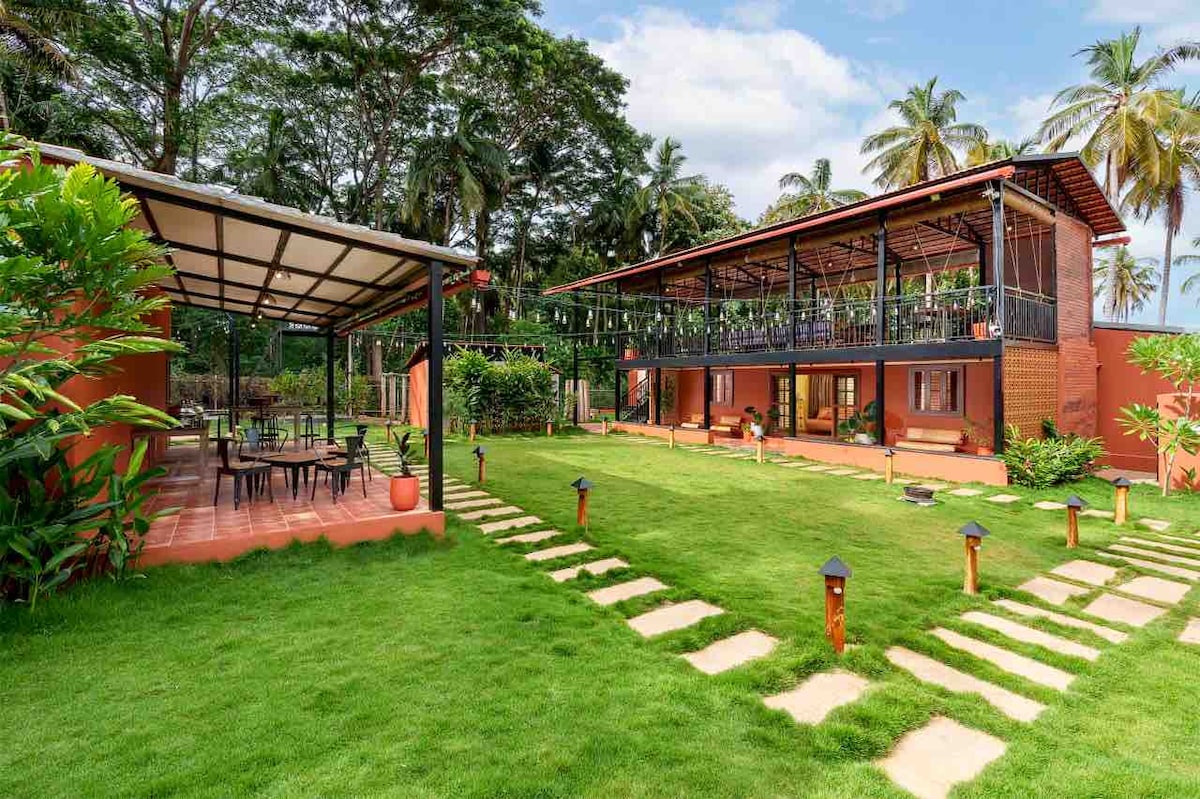 The outdoor area features a well-maintained lawn, bordered by tropical plants. A stone pathway leads to a spacious two-story farmhouse with a balcony overlooking the garden. An adjacent covered dining area includes several tables and chairs, ideal for outdoor gatherings.
