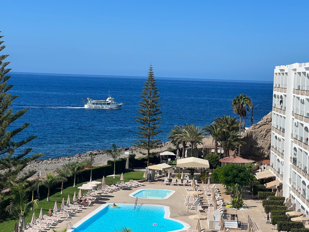 The image depicts a serene ocean view featuring a large swimming pool surrounded by lounge chairs and umbrellas. Palm trees provide shade, while a boat is seen sailing in the distance on calm waters. The clear blue sky enhances the peaceful atmosphere.