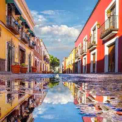 A vibrant street scene captures the colorful facades of buildings with balconies adorned with plants. A puddle reflects the sky and surrounding architecture, creating an engaging visual symmetry. The atmosphere conveys a welcoming charm in this historic area.