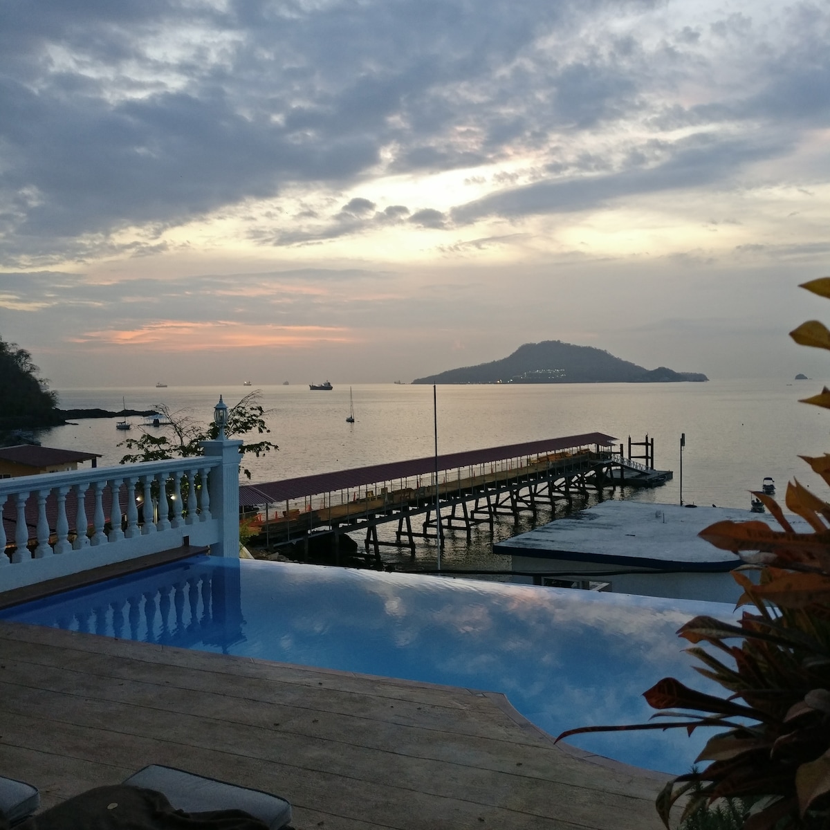An infinity pool stretches toward the horizon, reflecting soft sunset colors above a calm sea. A wooden pier extends into the water, leading to a distant island. Lush foliage frames the scene, providing a serene backdrop to this beachside view.