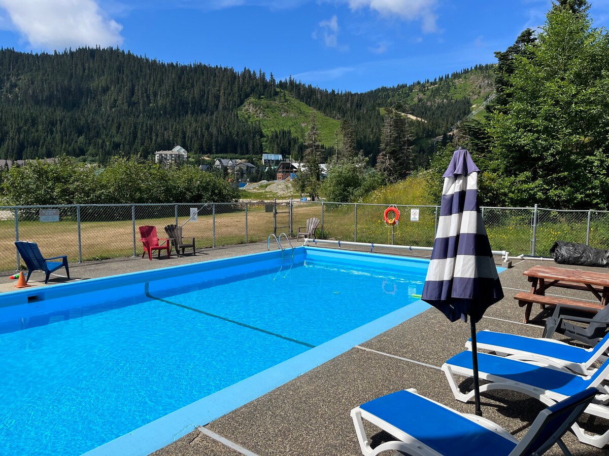 An outdoor pool is surrounded by a sun terrace featuring lounge chairs and umbrellas. Lush greenery and mountains are visible in the background, with a clear blue sky above. The pool area is enclosed by a chain-link fence, enhancing the sense of privacy.