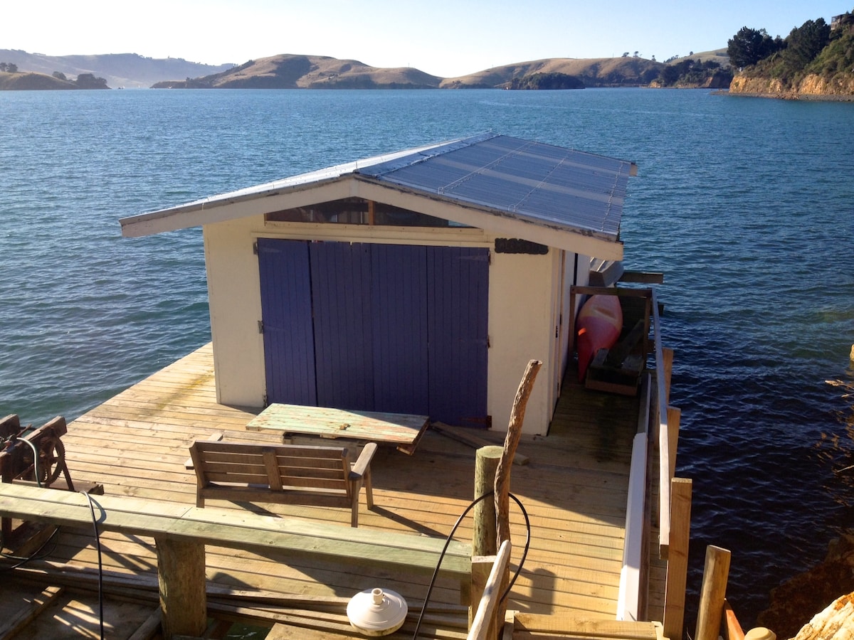 A self-contained guesthouse sits on a wooden deck overlooking tranquil waters. The structure features large double doors in a soft blue hue and solar panels on the roof, with a small bench positioned nearby. Scenic hills are visible in the background.