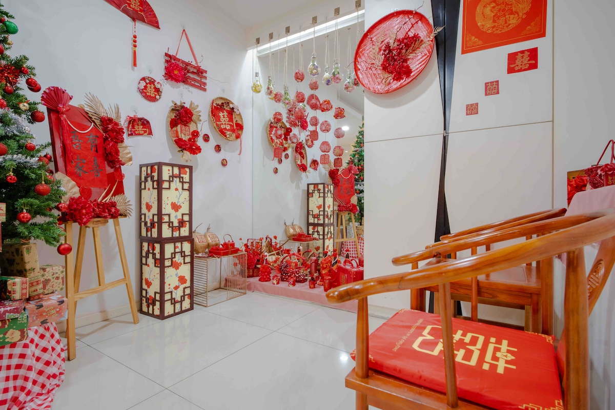 A festive corner showcases vibrant red decorations, including lanterns, floral arrangements, and traditional ornaments. A pair of wooden chairs is positioned nearby, adding an inviting touch. Gifts wrapped in decorative paper are placed strategically, enhancing the celebratory atmosphere.