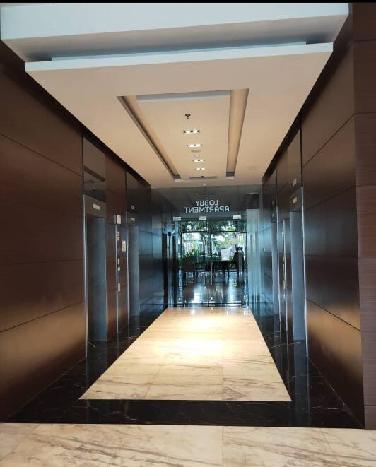 A modern corridor is depicted, featuring polished marble flooring and sleek wooden paneling. The ceiling is illuminated by recessed lighting, and glass doors at the end provide a view of a vibrant communal area. Elevator access is visible along the sides of the hallway.