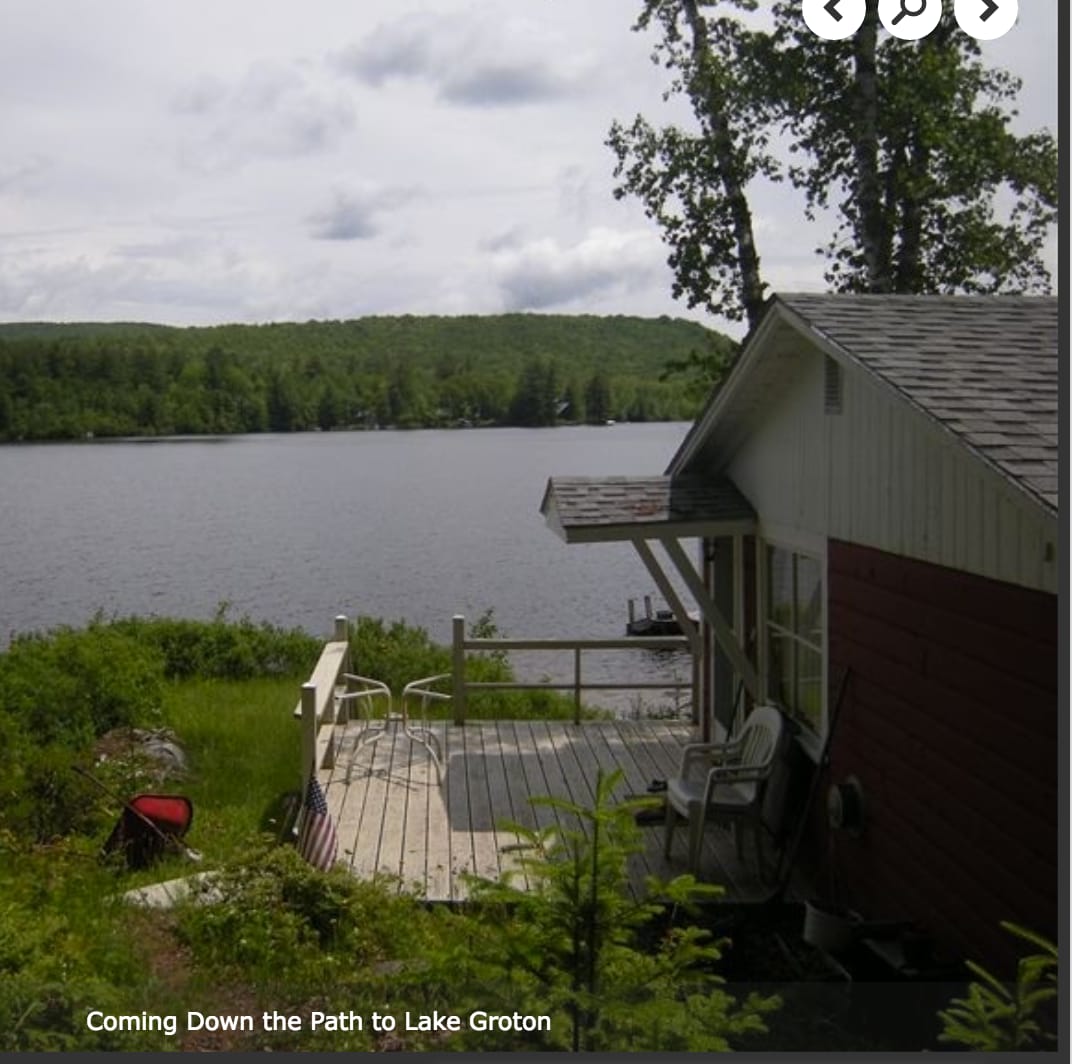 A clear view of the tranquil Lake Groton is visible from the wooden deck of the camp. A comfortable chair sits on the porch, with trees framing the peaceful landscape. The path leads down to the water, enhancing the camp's serene connection to nature.