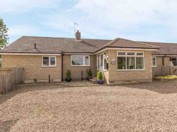 The exterior of the semi-detached cottage is showcased, featuring a stone facade and large windows that offer ample natural light. A gravel parking area is visible, along with a pathway leading to the entrance. Surrounding greenery adds to the peaceful setting.