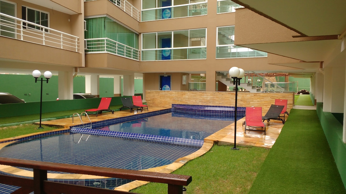 A serene pool area features a rectangular swimming pool surrounded by a grass lawn. Several lounge chairs in red and black are positioned along the poolside, and outdoor lighting fixtures provide illumination. The backdrop includes a modern multi-story building with large glass windows.