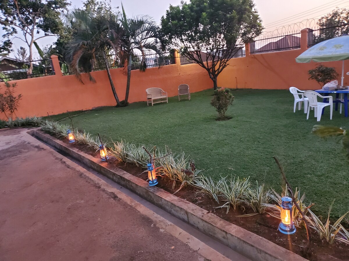 A well-maintained garden area is depicted, featuring a neatly manicured lawn bordered by low shrubs. Two wicker chairs are positioned under an umbrella near a table, while lanterns line the pathway, providing soft illumination. The garden is enclosed by a vibrant orange wall, creating a private space.