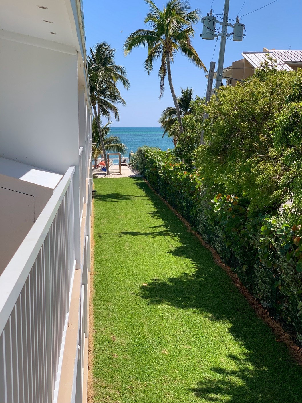 A lush green pathway leads from the property to the beach, framed by tall palm trees. The serene blue ocean is visible in the distance, beyond the neatly trimmed grass and vibrant foliage lining the path.