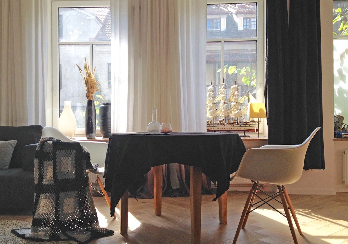 Natural light fills the living area, highlighting a dining table with a dark tablecloth and stylish chairs. Nearby, a cozy sofa and decorative elements, such as a vase and model ships on a shelf, add character to the space.