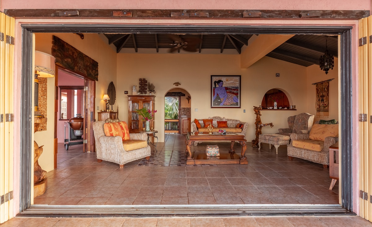 An open living area is framed by large sliding doors, allowing for a breezy connection to the outdoors. The space features cozy seating arrangements, warm-colored textiles, and distinctive artwork on the walls, enhancing the relaxed atmosphere of the villa.