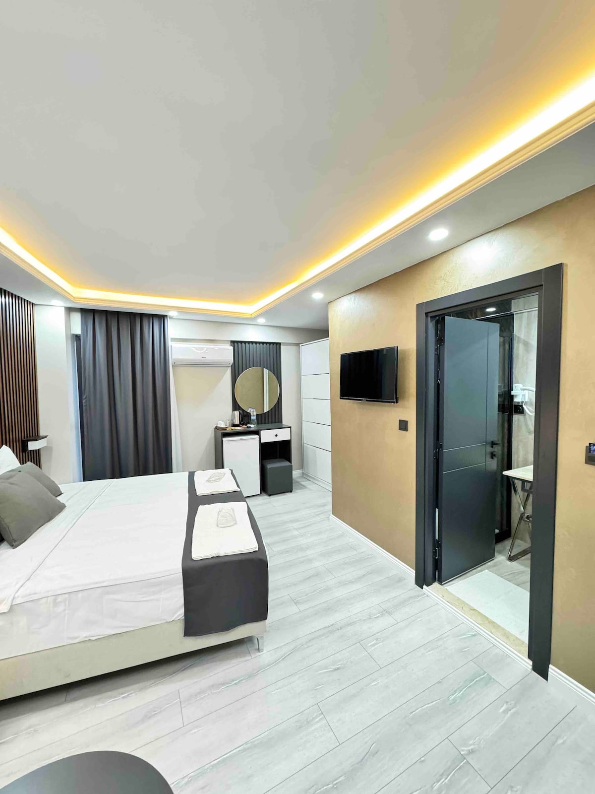 A modern room features a spacious bed with neutral bedding and accent pillows. Soft lighting highlights the sleek design. A wall-mounted television is positioned in front, and a bathroom door is visible on the right, with a mirror and dresser adding functionality to the space.