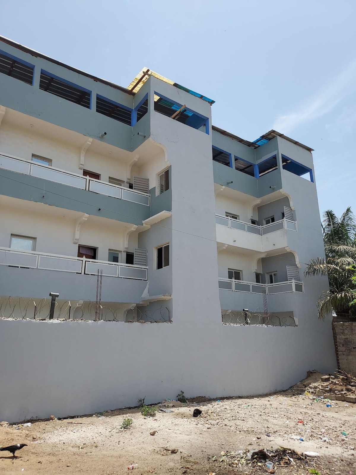 A modern three-story building is depicted, featuring a light grey exterior with balconies on each level. Reinforced by blue accents on the upper section, the structure complements its surroundings, which include greenery and a clear blue sky overhead.