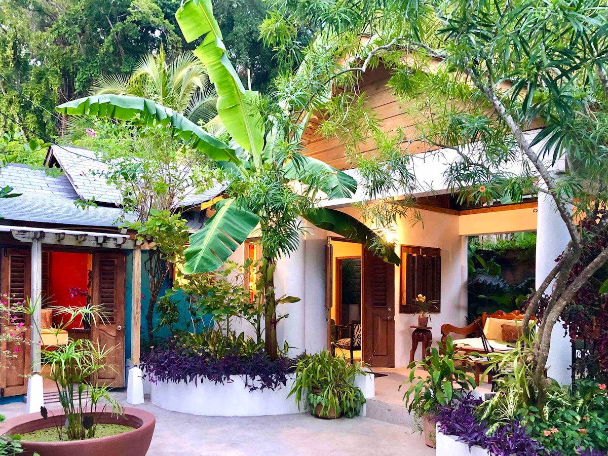 A charming cottage is framed by lush greenery and tropical plants. Entryways are accentuated by wooden shutters, and the inviting porch leads to a cozy seating area. The vibrant colors of the surrounding foliage blend harmoniously with the cottage's architecture, creating a serene outdoor ambiance.