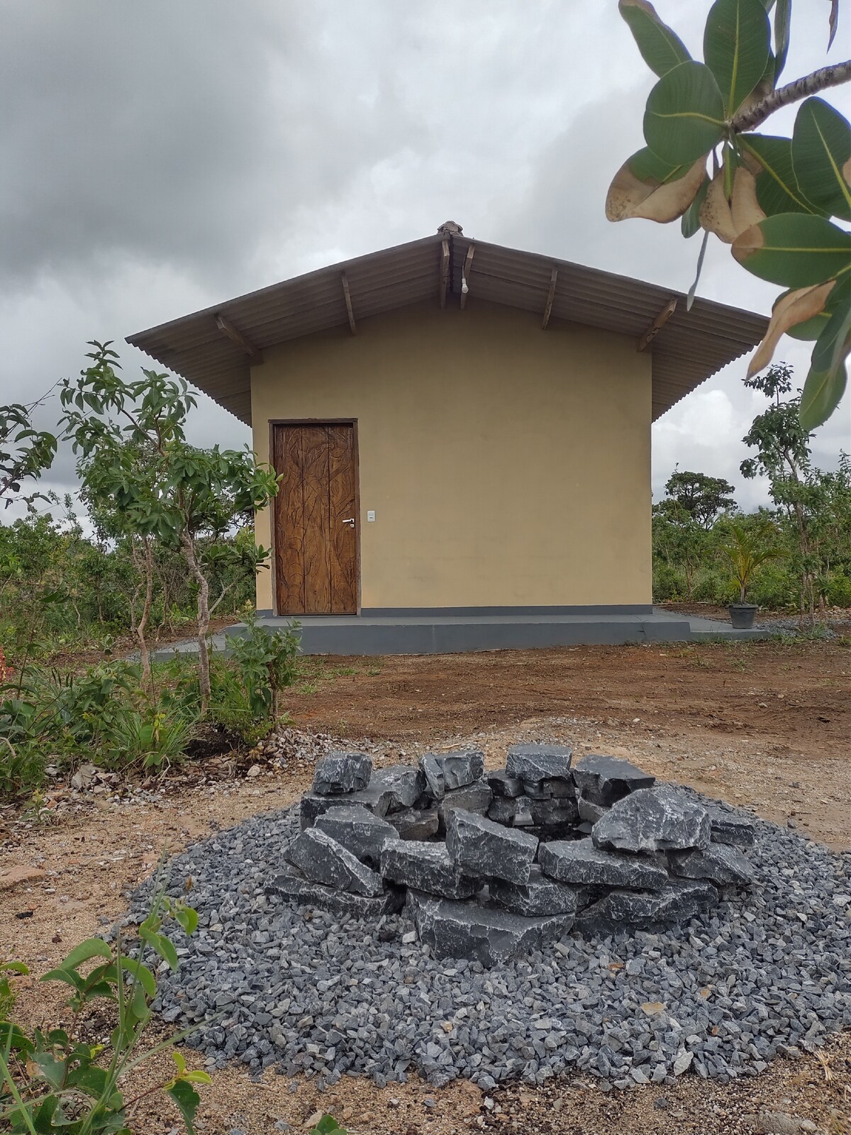 A simple, single-story building is situated within a natural landscape, featuring a wooden door and a thatched roof. A stone fire pit is centrally placed, surrounded by gravel and vegetation, providing a welcoming outdoor space.