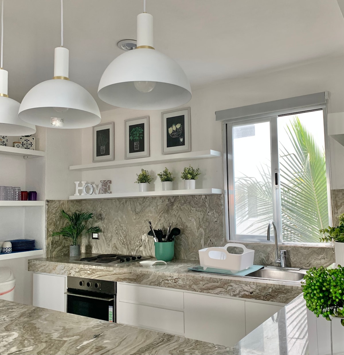 The kitchen features modern white cabinetry and a stylish marble countertop. Task lighting is provided by three pendant lights above the counter. A window allows natural light to illuminate the space, with a view of greenery visible through the glass.