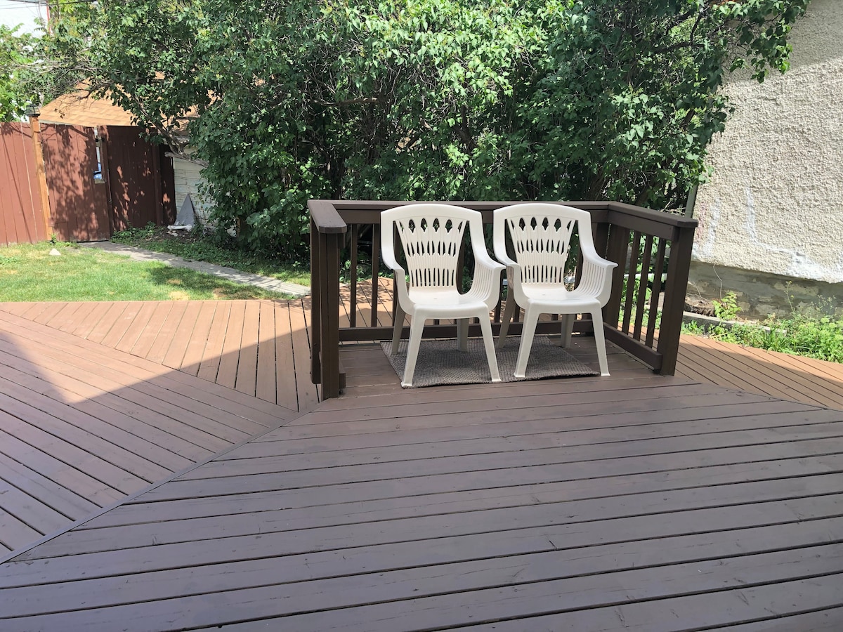 An outdoor deck features two white plastic chairs positioned near a low railing, providing a view of the surrounding greenery. The wooden surface of the deck showcases a rich brown color, creating a spacious area for relaxation.