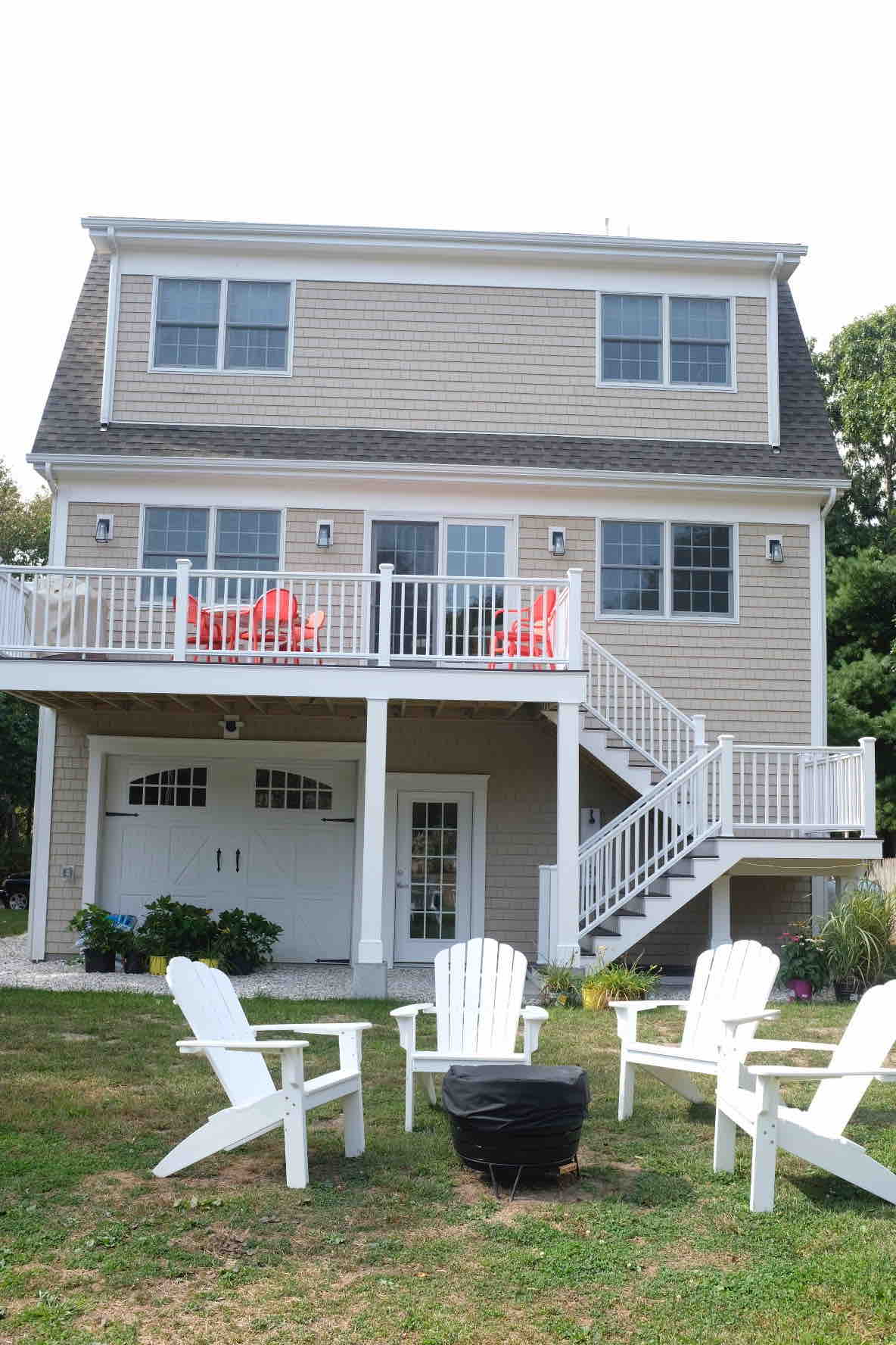 A three-story beach house is presented, featuring a light-colored exterior. A deck with two bright chairs overlooks the yard. White adirondack chairs are arranged around a fire pit in the grassy area, creating an inviting outdoor space. A staircase leads to the upper floors.
