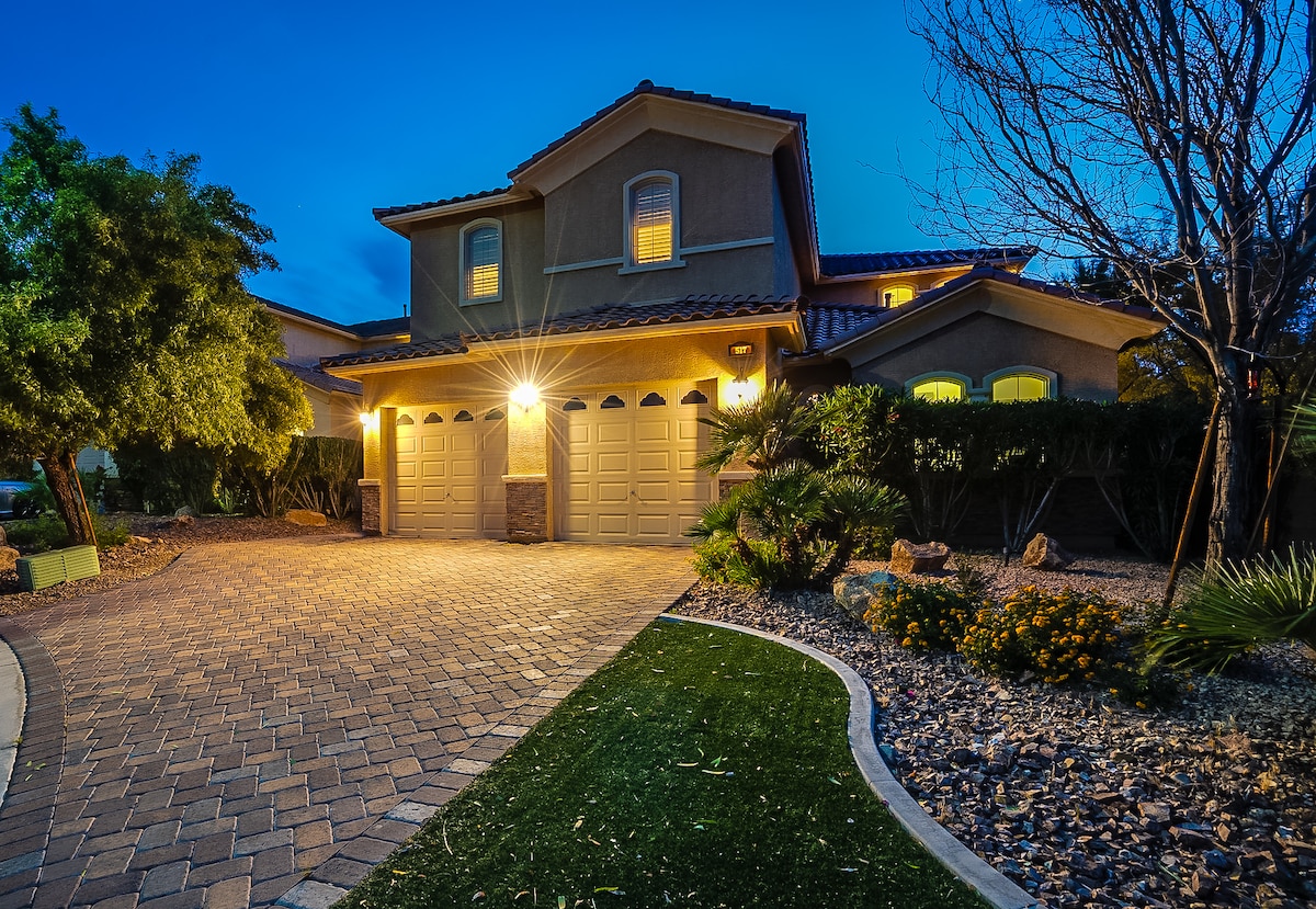 A two-story home is displayed, showcasing a paved driveway leading to a three-car garage. Subtle exterior lighting highlights the entryway, while surrounding landscaping features manicured grass and various shrubs, creating a welcoming curb appeal. Soft evening light enhances the home's exterior features.