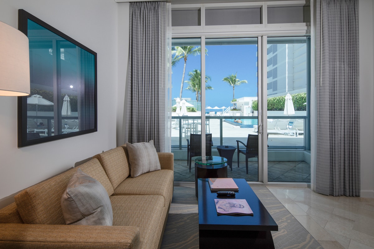 A comfortable seating area is presented with a neutral-toned sofa and a sleek coffee table. Glass doors offer a view of the outdoor space, revealing poolside amenities and palm trees. Natural light fills the room, enhanced by light grey curtains framing the doors.