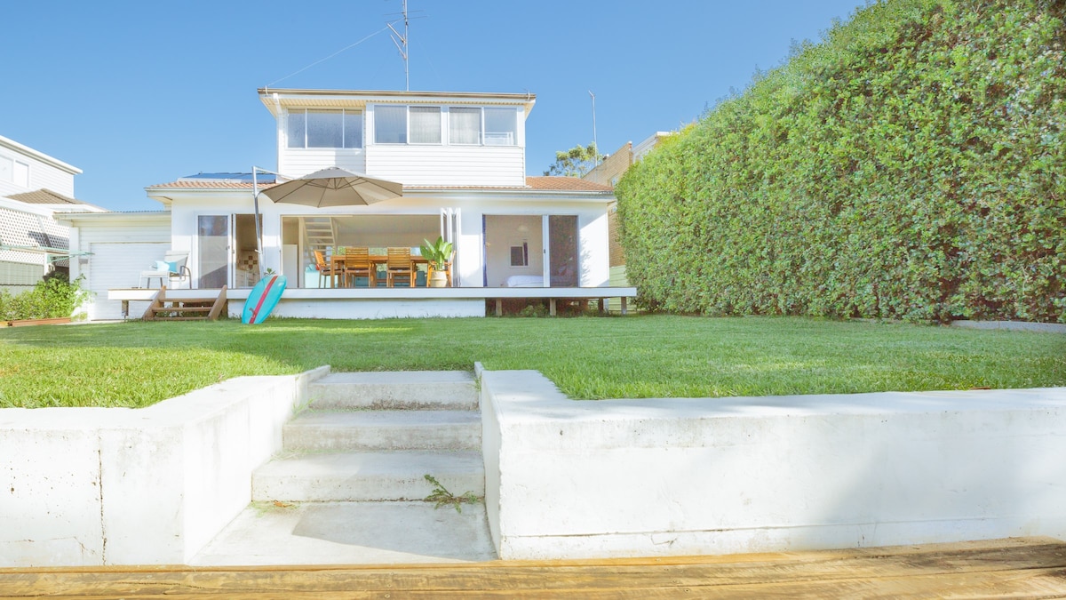 The house features a well-maintained lawn leading up to a spacious deck. An outdoor dining area, shaded by an umbrella, is visible alongside colorful lounge chairs. A clean exterior with large windows and a second-story balcony opens to the backyard, creating a welcoming outdoor space.