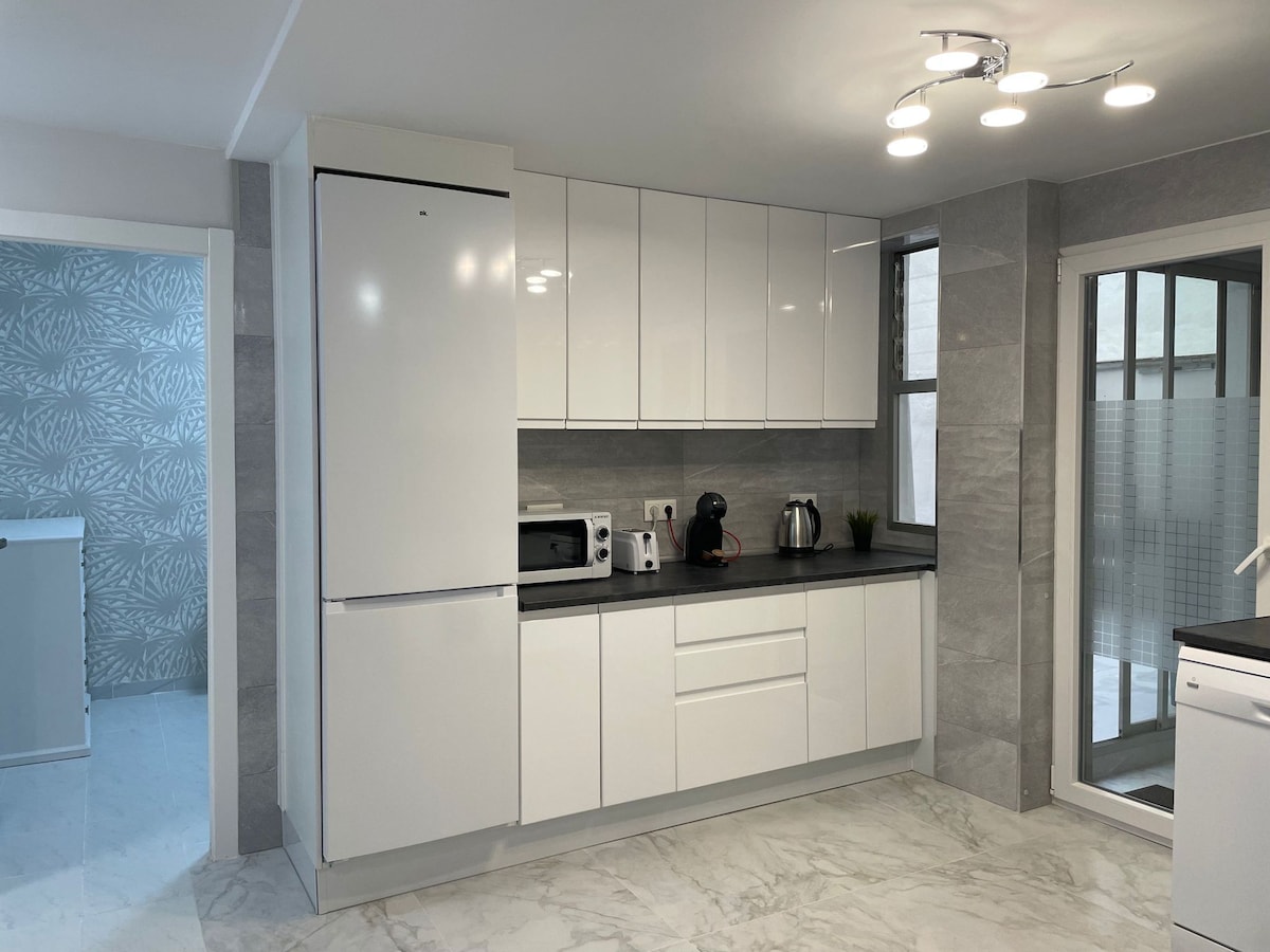 A modern kitchen features sleek, white cabinetry and a black countertop. Essential appliances include a refrigerator, microwave, and kettle. Natural light enters through a nearby window, enhancing the minimalist design. A doorway leads to an adjoining room, adding to the kitchen's spacious feel.