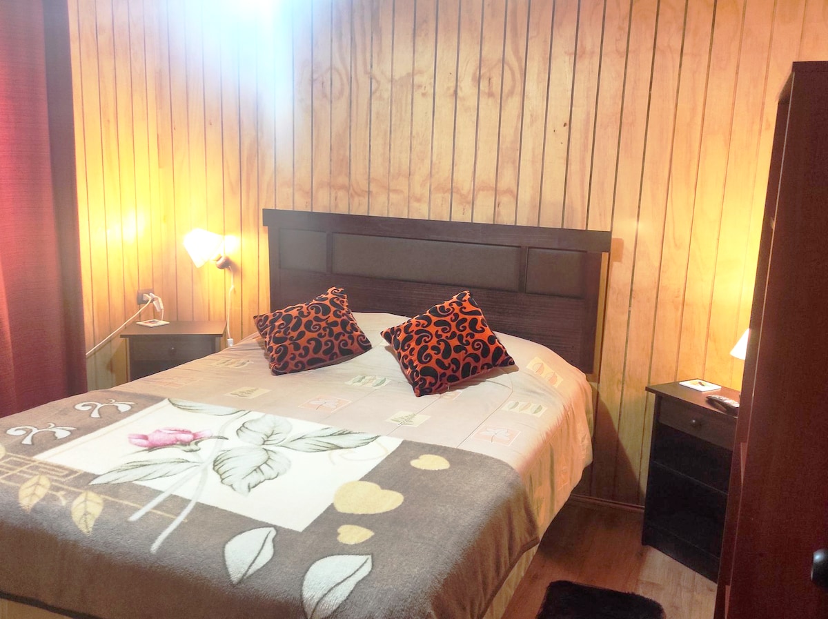 A cozy sleeping area is presented with a spacious bed dressed in neutral tones. Two decorative cushions with bold patterns are placed at the head of the bed. Wooden paneling and soft lighting contribute to a warm ambiance.