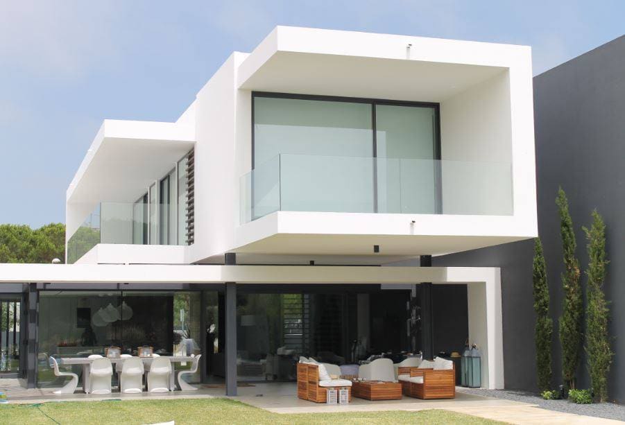 The modern villa showcases a minimalist exterior with large glass windows and a balcony on the upper level. A spacious outdoor area features comfortable seating arrangements and a large dining table, blending indoor and outdoor living. Lush greenery surrounds the property.