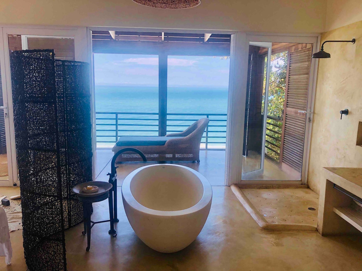 A spacious bathroom features a freestanding tub, paired with a wooden shower area. Large glass doors frame stunning ocean views, while a comfortable lounge chair is positioned nearby. A decorative privacy screen adds texture to the open space, enhancing the serene atmosphere.