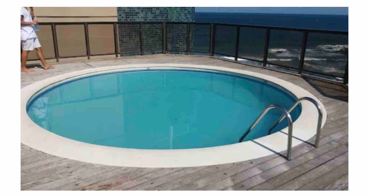 A circular pool is situated on a wooden deck, featuring a clear blue water surface. Two metal handrails provide access, and the panoramic ocean view is visible beyond the railing, highlighting the natural surroundings.
